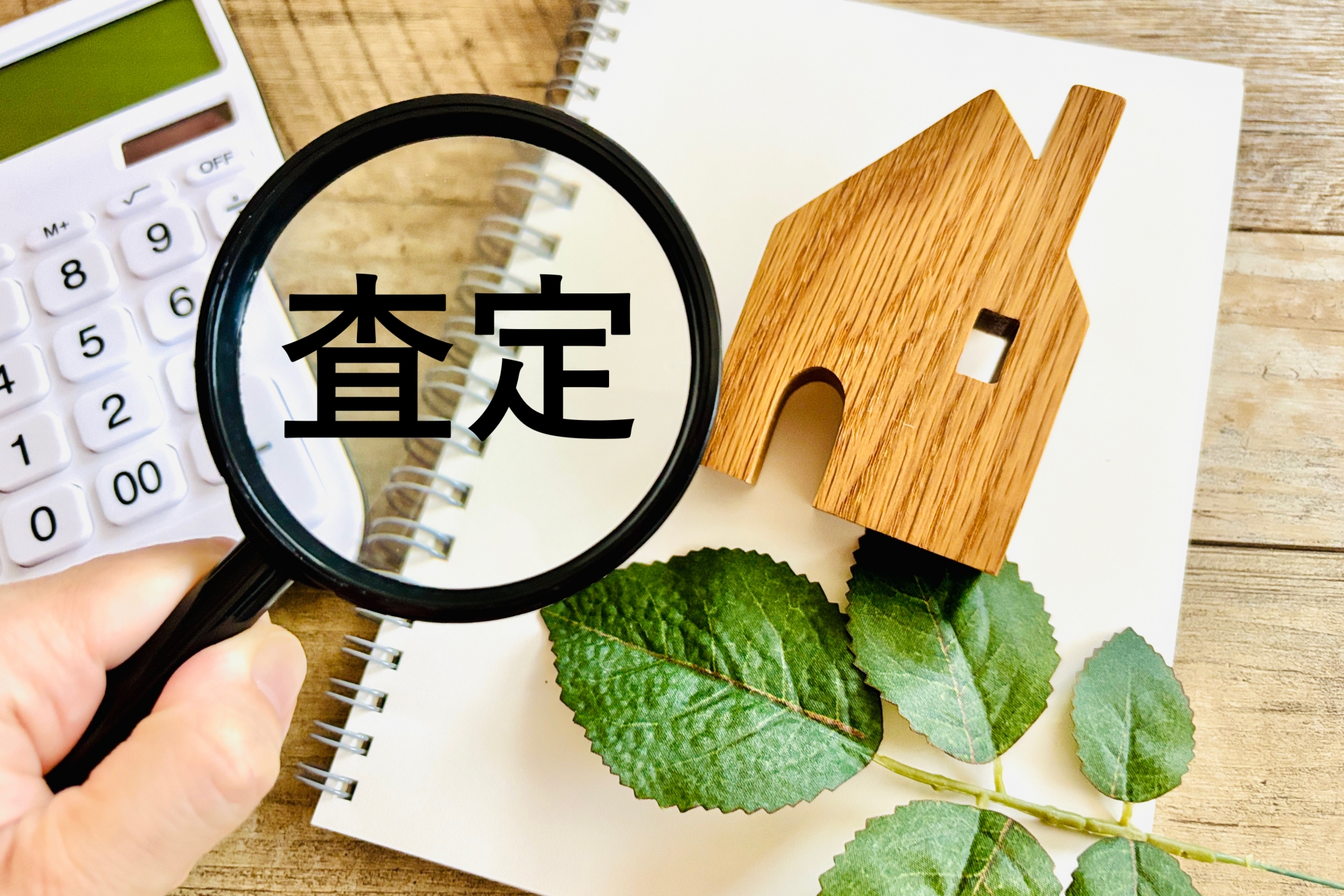 不動産売却のための査定を虫眼鏡で確認する様子、電卓と木の家モデル